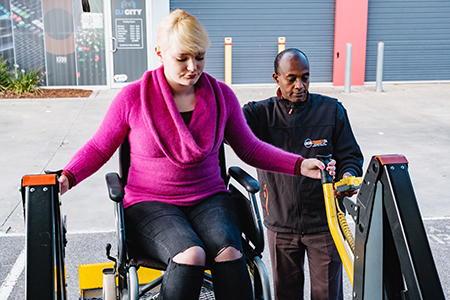 A picture containing a woman in a wheelchair and a man pulling a lever to lift the wheelchair up into a wheelchair accessible van.